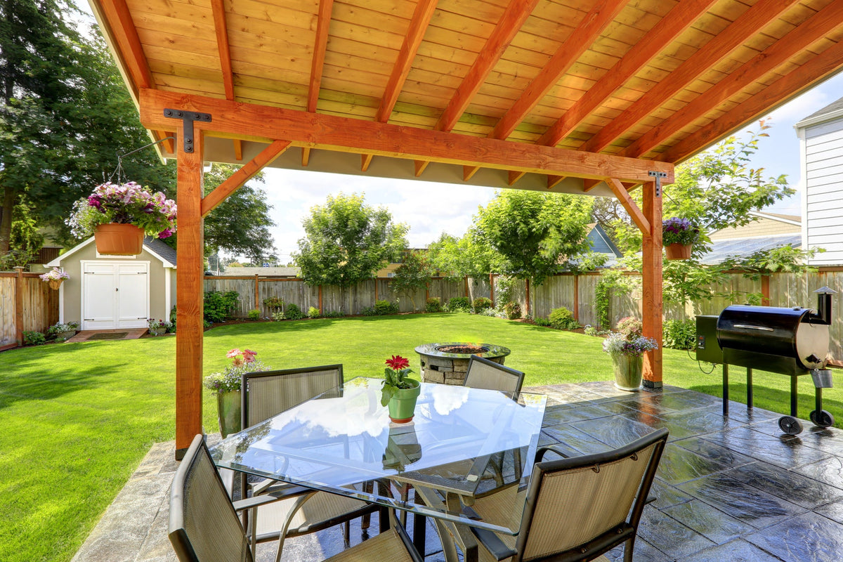 Gartenpavillion selber bauen – Blick unter einem hölzernen Pavillondach auf einen gedeckten Terrassentisch, Grill und eine gepflegte Rasenfläche mit Bäumen und Blumen.